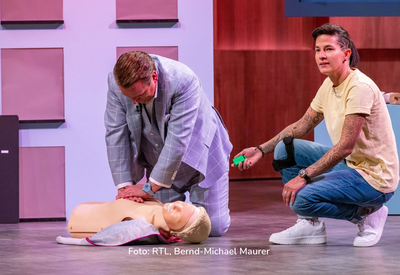 TV-/Bühnenfoto: Person in Anzug führt Herzdruckmassage an einer Übungspuppe aus; daneben kniet eine weitere Person; Text unten: „Foto: RTL, Bernd-Michael Maurer“.
