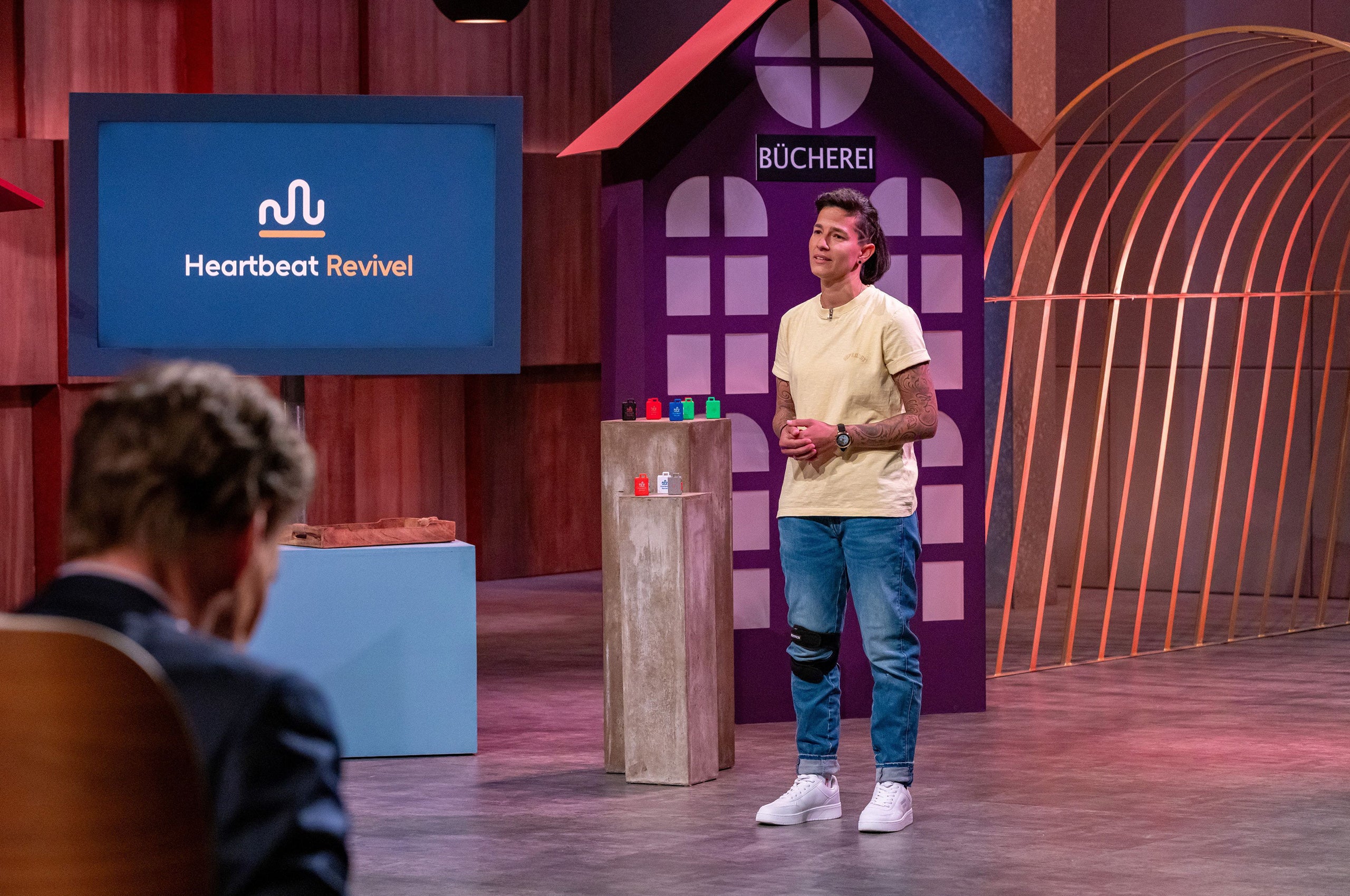 TV-/Bühnenfoto: Person steht im Studio vor Bildschirm mit „Heartbeat Revival“-Logo; im Vordergrund unscharf eine sitzende Person.